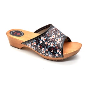 May include: Black and white floral print leather clog with a wooden heel and sole. The clog has a small opening at the toe and a black leather strap across the top. The sole has a textured pattern. The clog has a red star logo on the side with the text "Original Leather Austria"