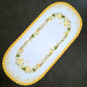 May include: A white oval table runner with a yellow scalloped border. The runner features a floral design with yellow and green daisies and small yellow birds.