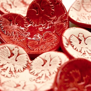 May include: A collection of red and white ceramic dishes with a Polish eagle emblem. The dishes are heart-shaped and round, with a glossy finish. The eagle design is embossed on the surface, creating a textured effect.