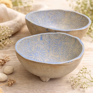 May include: Two handmade ceramic bowls with an irregular, organic shape and a light beige, speckled finish. The interior of each bowl features a stamped, repeating floral pattern in a soft blue glaze, with blue detailing along the rim.
