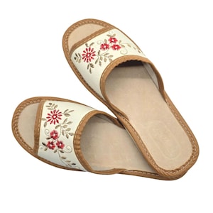 May include: A pair of open-toe slippers featuring a cream-colored upper band with red floral embroidery and green stems. The slippers have a tan-colored trim along the edges and a light beige footbed, providing a simple and functional design.
