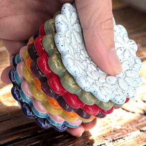 May include: A stack of circular ceramic coasters featuring a raised floral pattern. The top coaster is white with black speckles, followed by layers in shades of green, red, dark purple, orange, yellow, pink, light blue, and dark blue.