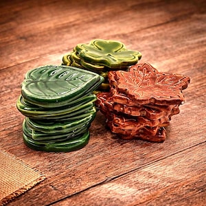 May include: A collection of ceramic leaf-shaped coasters stacked in three distinct piles. One stack features dark green heart-shaped leaves, another consists of green veined leaves, and the third pile contains brown maple-shaped leaves with detailed textures.
