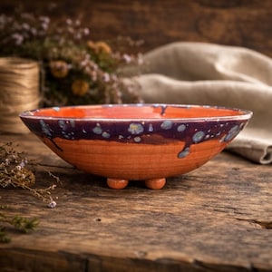 May include: A shallow ceramic bowl with three small feet, featuring a terracotta-colored base and a dark purple glazed rim with light blue speckled accents that drip down the sides, creating a distinct contrast between the matte and glossy textures.