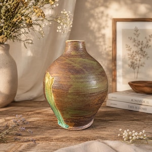May include: A round ceramic vase with a narrow neck, featuring a textured surface with mottled brown and green glaze. The vessel has a wide, bulbous body that tapers toward the top opening, showcasing organic color variations and a rustic finish.