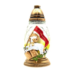 May include: A glass votive candle holder with a gold metal base and lid. The holder features a white lamb with a red flag and a gold halo, surrounded by green leaves and white flowers. The words "A.D." are visible on the base.