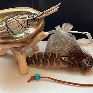 May include: A white sage smudge stick in a seashell holder with a wooden stand, a small bag of dried herbs, and a feather smudge fan with a turquoise bead and brown leather cord.