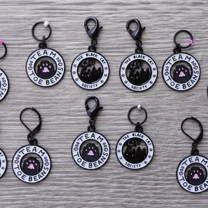 May include: Twelve black metal stitch markers with white lettering. Six markers say "Team Toe Beans" with a pink paw print. Six markers say "The Black Cat Society" with a black cat silhouette.