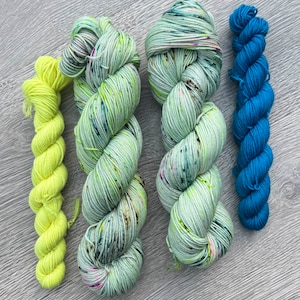 May include: Four skeins of yarn in different colors: a bright yellow, a light green with speckles of color, a darker green with speckles of color, and a deep blue.