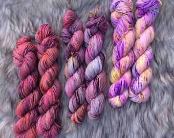 May include: Six skeins of hand-dyed yarn in shades of pink, purple, and yellow. The yarn is arranged on a gray, faux fur surface. The colors blend together, creating a variegated effect, perfect for knitting or crocheting projects.