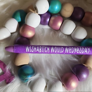 May include: Purple glitter pen with the text "Wishabitch Would Wednesday" in white lettering. The pen is surrounded by a garland of wooden beads in various colors, including white, purple, teal, and pink.