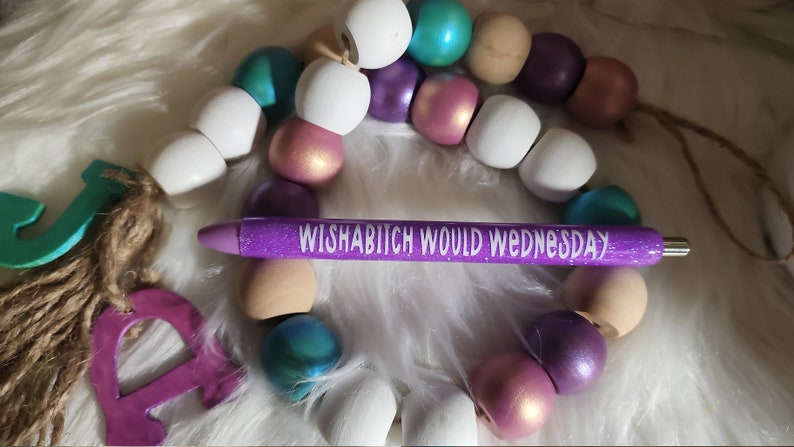 May include: Purple glitter pen with the text "Wishabitch Would Wednesday" in white lettering. The pen is surrounded by a garland of wooden beads in various colors, including white, purple, teal, and pink.
