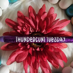May include: Purple glitter pen with white text that reads "THUNDERCU*T TUESDAY". The pen is lying on a white fuzzy surface with a red flower.
