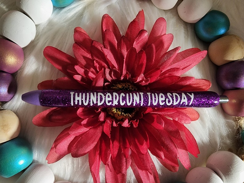May include: Purple glitter pen with white text that reads "THUNDERCU*T TUESDAY". The pen is lying on a white fuzzy surface with a red flower.
