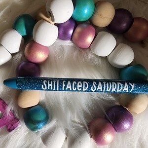 May include: A blue glitter pen with the text "Shit Faced Saturday" written in white letters. The pen is lying on a white fuzzy surface with a string of colorful wooden beads.