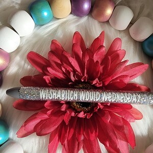 May include: A silver glitter pen with the text "WISHABITCH WOULD WEDNESDAY" printed in white. The pen is lying on a red flower with a white background.