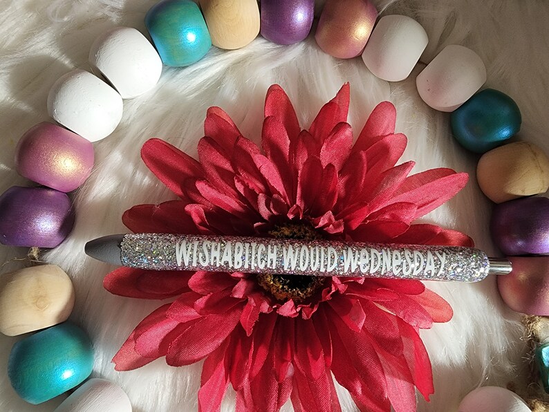 May include: A silver glitter pen with the text "WISHABITCH WOULD WEDNESDAY" printed in white. The pen is lying on a red flower with a white background.