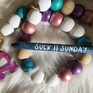 May include: A blue glitter pen with the text "SUCK IT SUNDAY" written in white. The pen is lying on a white fuzzy surface with a string of colorful wooden beads.