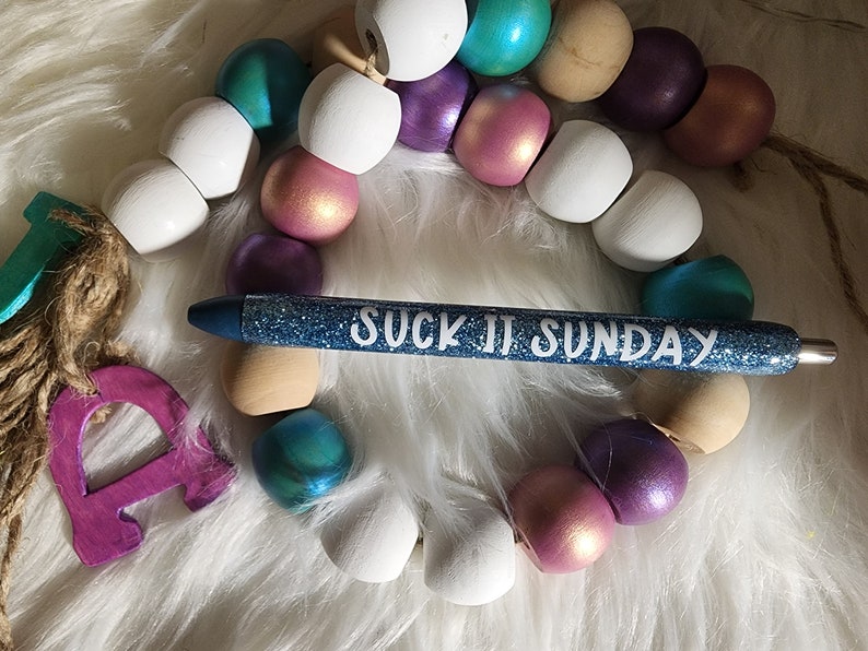 May include: A blue glitter pen with the text "SUCK IT SUNDAY" written in white. The pen is lying on a white fuzzy surface with a string of colorful wooden beads.