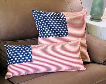 Könnte beinhalten: Zwei dekorative Kissen mit amerikanischem Flaggenmuster. Die Kissen liegen auf einem braunen Ledersofa. Das obere Kissen hat ein blaues Feld mit Sternen und roten und weißen Streifen. Das untere Kissen hat ein rot-weiß gestreiftes Design mit einem blauen Feld mit Sternen.