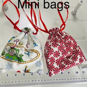 May include: Two mini drawstring bags, one with a white background and a colorful cartoon design, and the other with a white background and a red and white candy cane pattern. The bags are both tied with red ribbon.