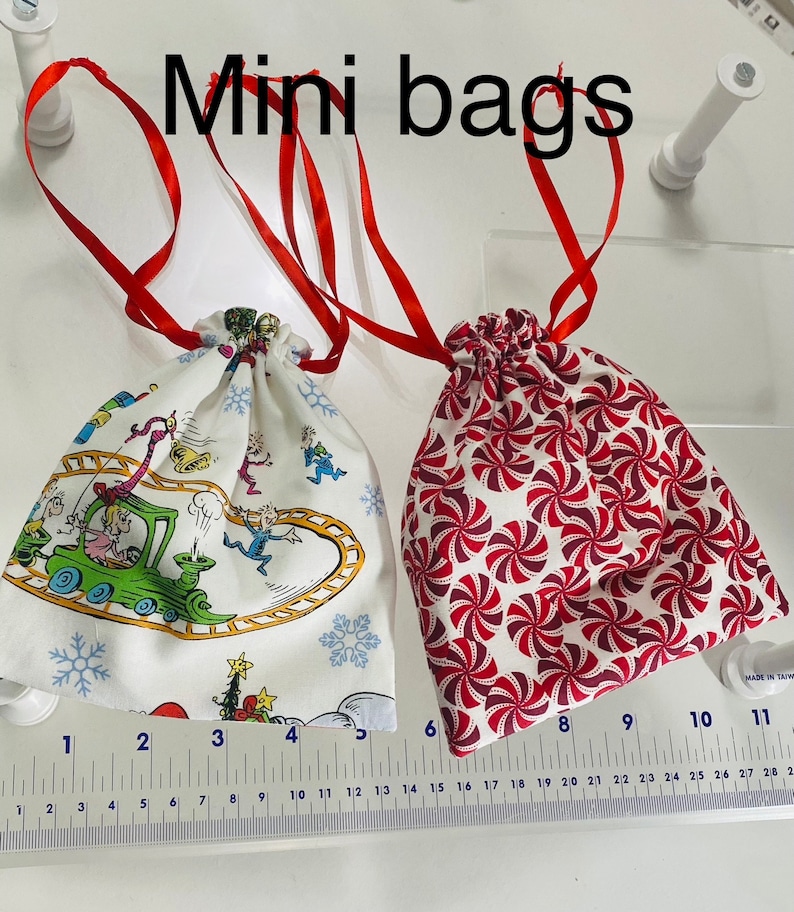 May include: Two mini drawstring bags, one with a white background and a colorful cartoon design, and the other with a white background and a red and white candy cane pattern. The bags are both tied with red ribbon.