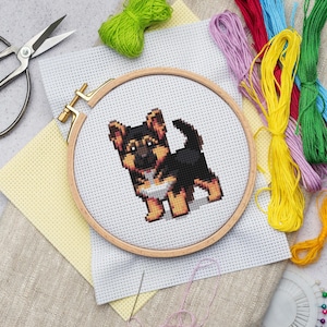 Puede incluir: Un proyecto de bordado de punto de cruz con un diseño de cachorro de pastor alemán en un bastidor de madera. El cachorro es negro, marrón y canela. Hilos de bordar coloridos, tijeras y tela están dispuestos alrededor del bastidor, con un tema artesanal.