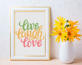 May include: A white frame with a gold trim surrounds a cross-stitch pattern that reads "live laugh love" in green, orange, and pink lettering. The frame is sitting on a wooden surface in front of a white wall with a vase of yellow flowers.