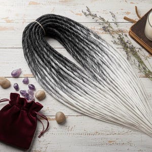 Puede incluir: Dreadlocks ombre en blanco y negro, atados con una banda. Una pequeña bolsa de terciopelo burdeos, cristales de amatista y piedras lisas están dispuestas alrededor del cabello. También se ve una vela.