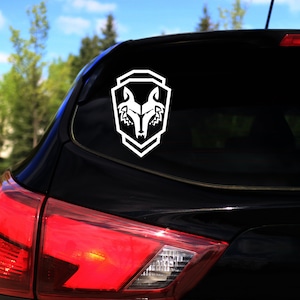 以下が含まれることがあります： 黒い車のリアウィンドウに、盾の形をした白いビニールステッカーに狼の頭が描かれている。