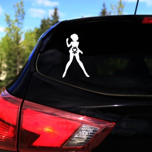 Op de afbeelding: Een zwarte auto met een witte silhouette-sticker van een vrouwelijke figuur met een symbool op haar borst. De sticker zit op de achterruit van de auto.