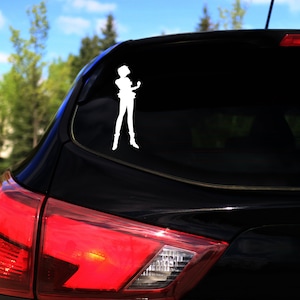 Op de afbeelding: Een witte vinyl sticker van een vrouwelijk anime-personage met kort haar, een kort rokje en laarzen aan, staand met haar armen langs haar zijde. De sticker zit op de achterruit van een zwarte auto.