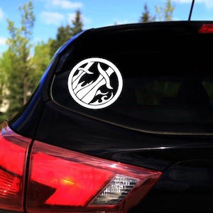 May include: A white vinyl decal of a stylized sword and flames design in a circle, applied to the back window of a black car.