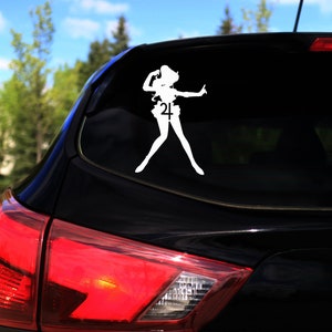 Op de afbeelding: Een zwarte auto met een witte vinyl sticker van een vrouwelijk anime-personage met een ster op haar borst en het nummer 24 op haar rok. De sticker zit op de achterruit van de auto.