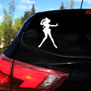 Op de afbeelding: Een wit silhouet van een vrouwelijke figuur in een rok en laarzen, met haar armen omhoog, is op de achterruit van een zwarte auto.
