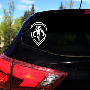 Puede incluir: Una calcomanía de vinilo blanco del símbolo mandaloriano en un coche negro. El símbolo es un escudo con una calavera y cuernos en su interior.