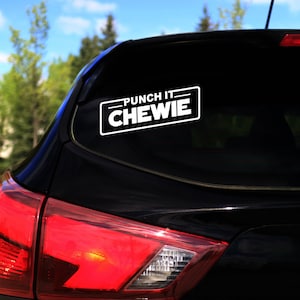 Puede incluir: Una pegatina rectangular blanca con el texto "PUNCH IT CHEWIE" en negrita, adherida a la ventana trasera de un coche negro. La pegatina está enmarcada por un borde negro. El fondo muestra árboles y un cielo azul.