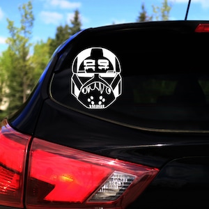 Könnte beinhalten: Weißes Vinyl-Aufkleber eines stilisierten Helms, wahrscheinlich für ein Fahrzeugfenster. Das Design zeigt einen detaillierten, kantigen Helm mit einem markanten Visier und Mundbereich. Der Aufkleber ist auf der Heckscheibe eines schwarzen Autos angebracht.