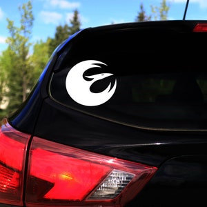 Könnte beinhalten: Weißer Vinyl-Autoaufkleber mit dem Symbol der Rebellen-Allianz, einem stilisierten Vogel in einem Kreis. Der Aufkleber befindet sich auf der Heckscheibe eines schwarzen Autos, wobei das Rücklicht sichtbar ist. Der Hintergrund zeigt Bäume und blauen Himmel.