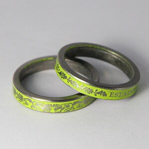 K&ouml;nnte beinhalten: Zwei silberne Ringe mit einer limegr&uuml;nen Emaille-Oberfl&auml;che. Die Ringe sind mit einem floralen Design und dem Wort "ESTADO" graviert.