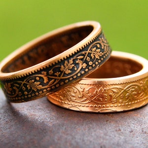 May include: Two gold-colored rings with intricate floral patterns. One ring has a black band with gold detailing, while the other is entirely gold. The rings are stacked, showcasing their detailed craftsmanship and design.