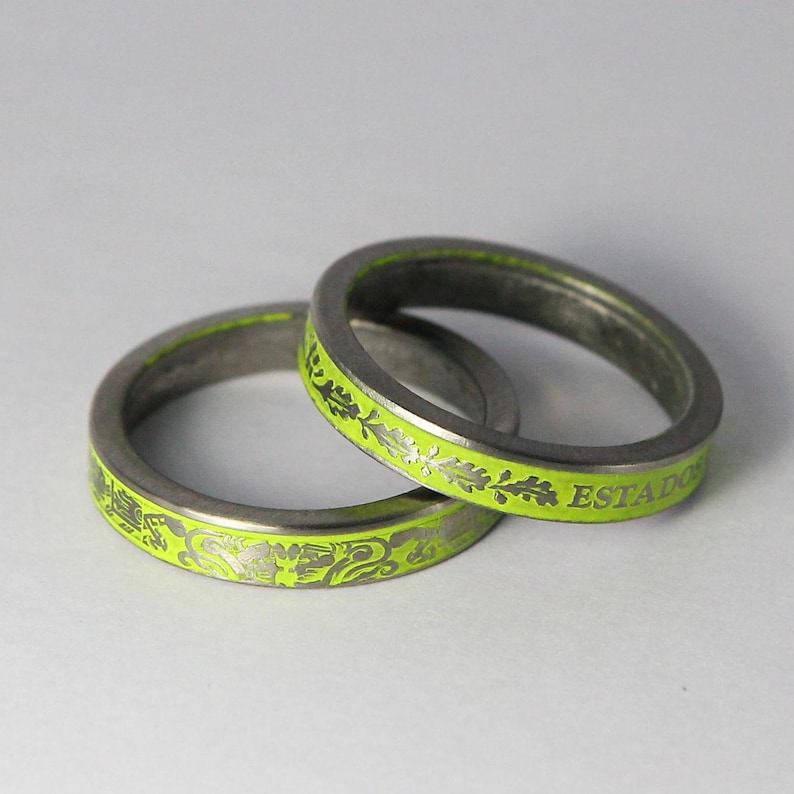 K&ouml;nnte beinhalten: Zwei silberne Ringe mit einer limegr&uuml;nen Emaille-Oberfl&auml;che. Die Ringe sind mit einem floralen Design und dem Wort "ESTADO" graviert.