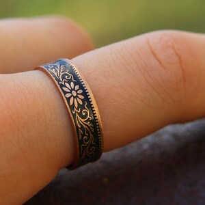May include: Close-up of a copper ring with a black enamel inlay. The ring features a floral and scroll design. The ring is worn on a finger. The ring's band is approximately 0.25 inches wide.