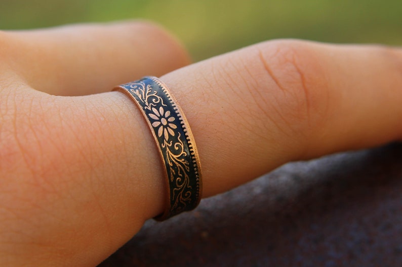 May include: Close-up of a copper ring with a black enamel inlay. The ring features a floral and scroll design. The ring is worn on a finger. The ring's band is approximately 0.25 inches wide.