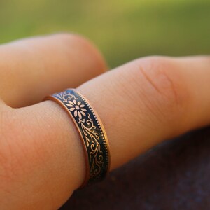 May include: A copper-colored ring with a dark blue enamel band featuring a floral and scroll design. The ring is worn on a finger, showcasing the intricate details of the decorative pattern. The ring's band is approximately 0.25 inches wide.
