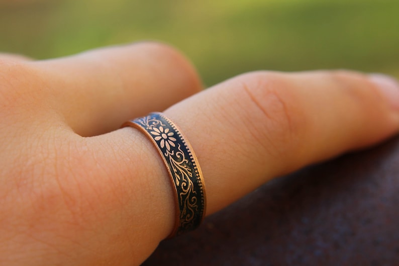 May include: A copper-colored ring with a dark blue enamel band featuring a floral and scroll design. The ring is worn on a finger, showcasing the intricate details of the decorative pattern. The ring's band is approximately 0.25 inches wide.