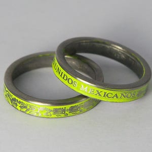 K&ouml;nnte beinhalten: Zwei silberne Ringe mit einer leuchtend gelben Inschrift. Die Inschrift auf einem Ring lautet "UNIDOS MEXICANOS". Der andere Ring hat ein dekoratives Design.