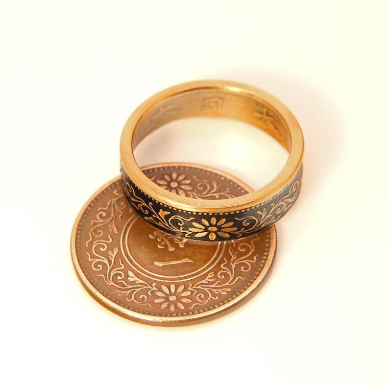 May include: A gold-toned ring with a black floral design sits atop a copper-colored coin. The ring's band features a repeating pattern of small flowers. The coin has an embossed floral design and the number "1".