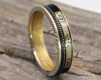 May include: A gold-toned ring with a black enamel band. The ring features etched detailing and a repeating pattern around the edges. The black band has etched text. The ring is set against a wooden surface.