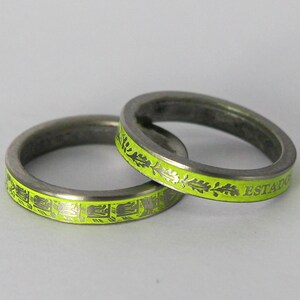 K&ouml;nnte beinhalten: Zwei silberne Ringe mit einer leuchtend gelben Emaille-Oberfl&auml;che. Ein Ring hat ein sich wiederholendes Muster aus stilisierten Bl&auml;ttern und dem Text "ESTADIO". Der andere Ring hat ein sich wiederholendes Muster aus stilisierten geometrischen Formen.